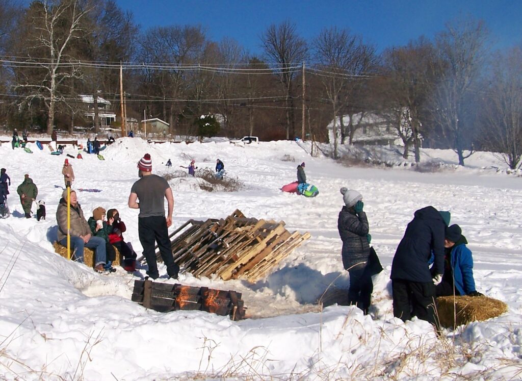 People sit around a warm bonfire. The fire tender is wearing a t-shirt (but still has a warm hat on).