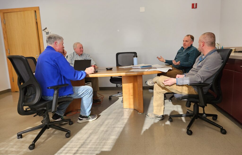 Four men sit in office-style chairs on two sides of a wooden table, gesturing and apparently deep in conversation. The chair at the head of the table is empty.