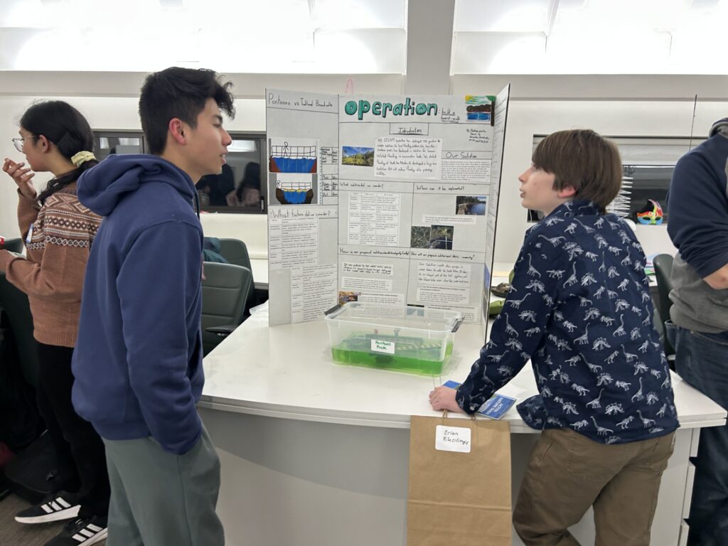 A high school aged boy talks with a junior high school aged boy. They stand on either side of a trifold display; in front of the trifold is a plastic container containing green-dyed water with a model of a pontoon bridge.
