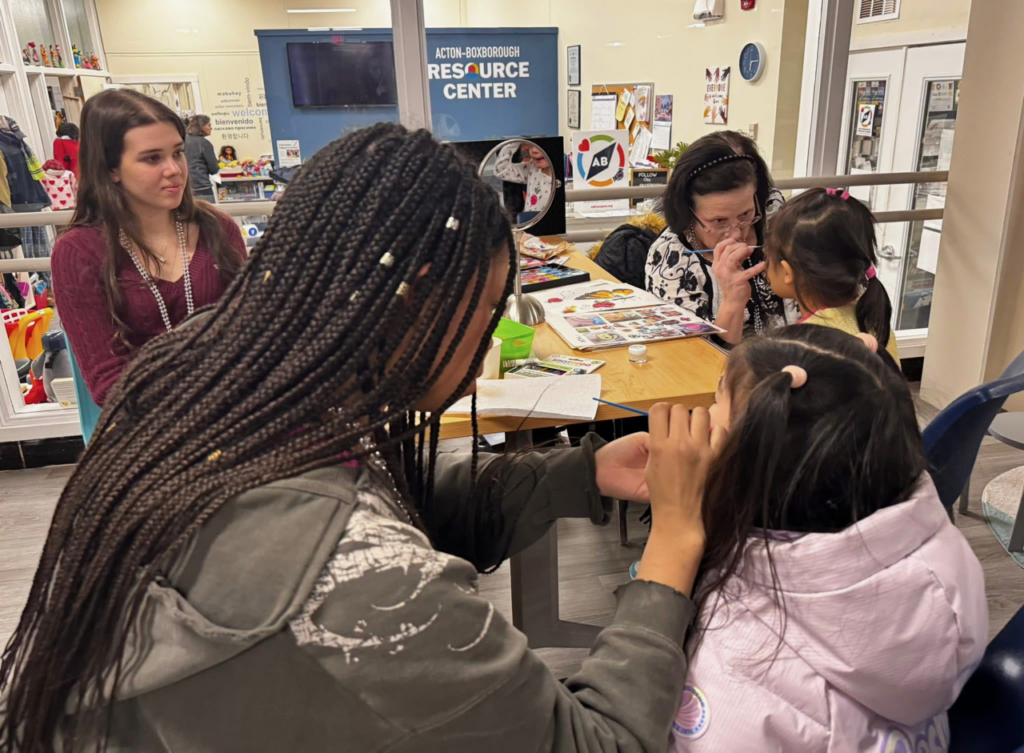 Two adult women carefully and delicately paint designs onto the faces of two young girls.