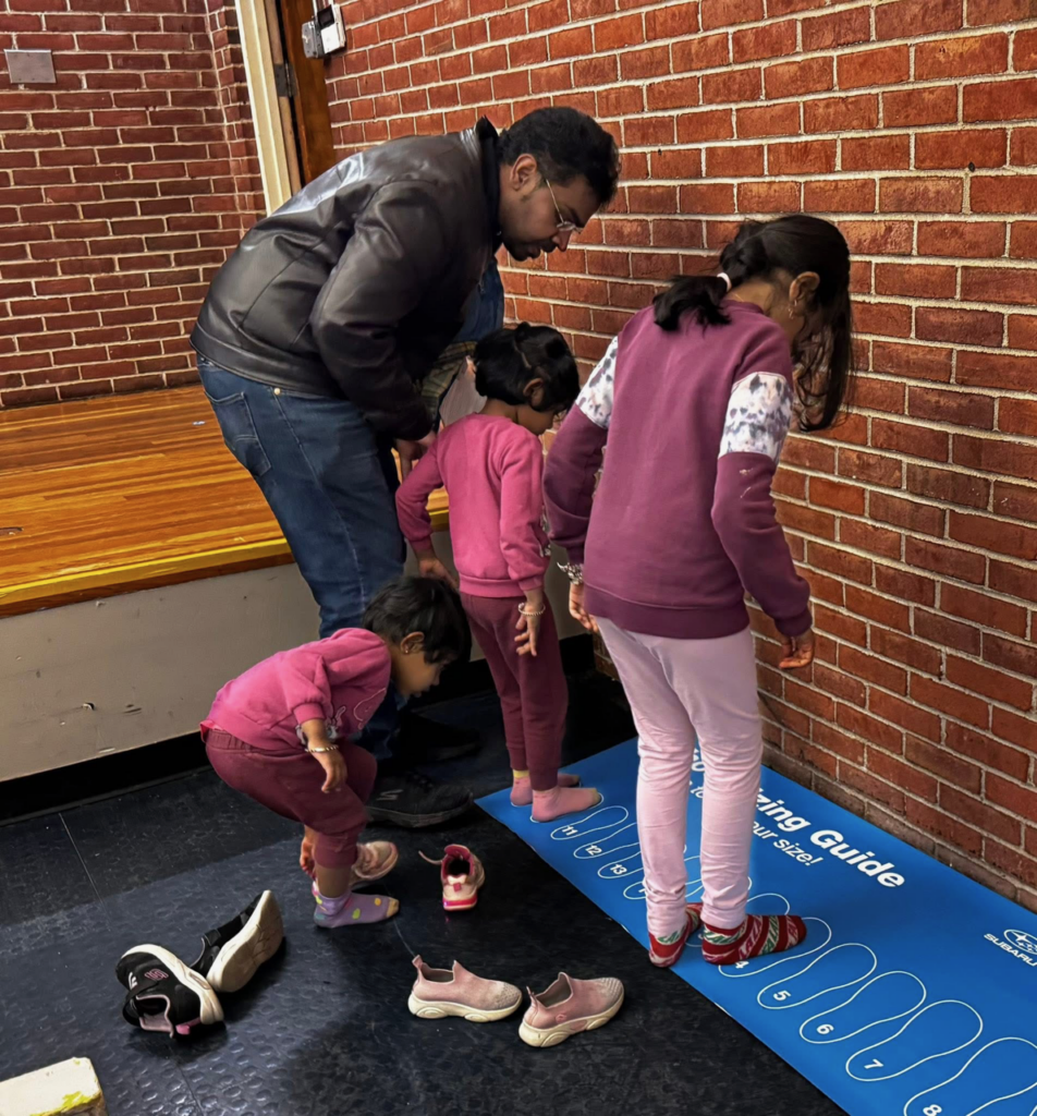 Two children, one large, one small, measure the size of their sock-clad feet on a mat printed with outlines of feet of different shoe sizes. These children's own shoes lie discarded on the floor. A younger child takes off her shoes in readiness for her own chance on the mat. An adult watches the process closely to see if help is needed.