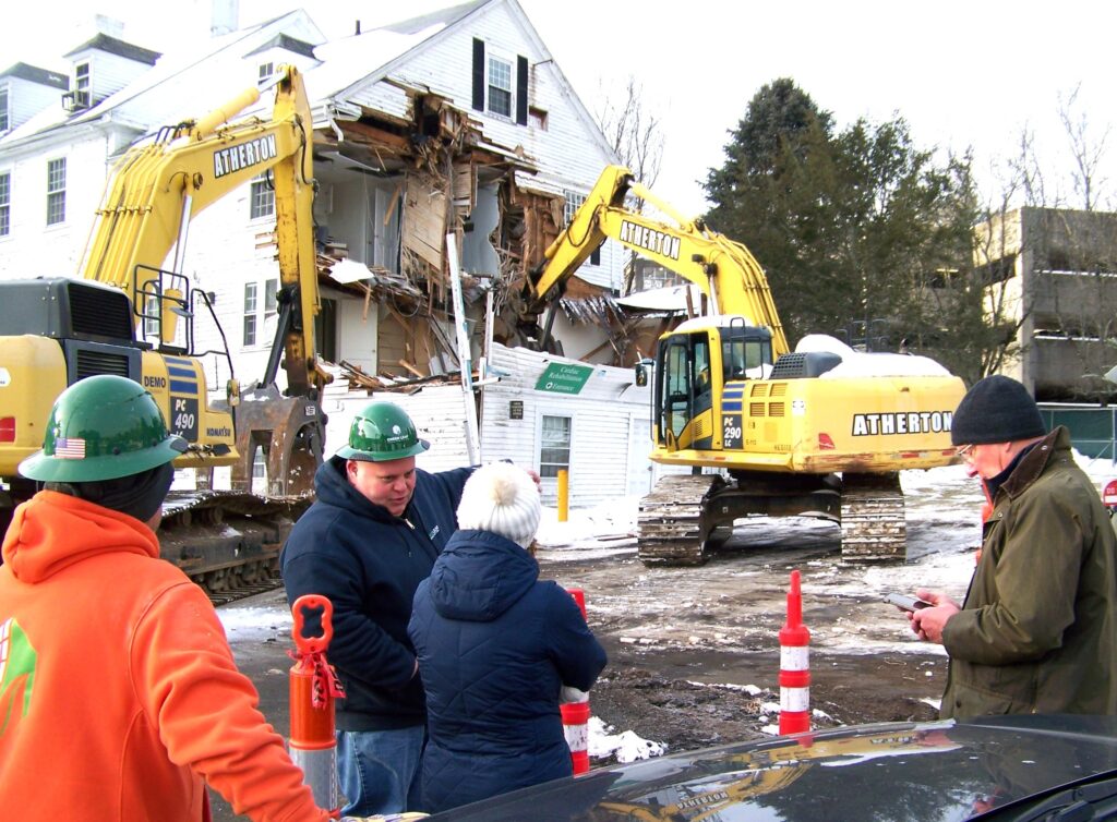 In the foreground, two people are chatting and a few other people are standing around. In the background, excavators are taking down the walls of a white house.