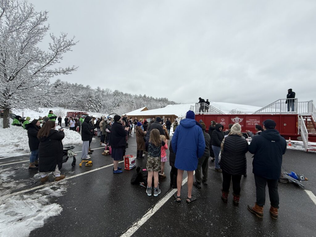 A group of people stand in front of the mobile plunge pool. At the very back, a man in a bright blue parka stands with bare legs and sandals.