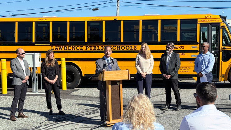Six people stand in front of a Lawrence Public Schools bus. One man in a suit is speaking at a podium.