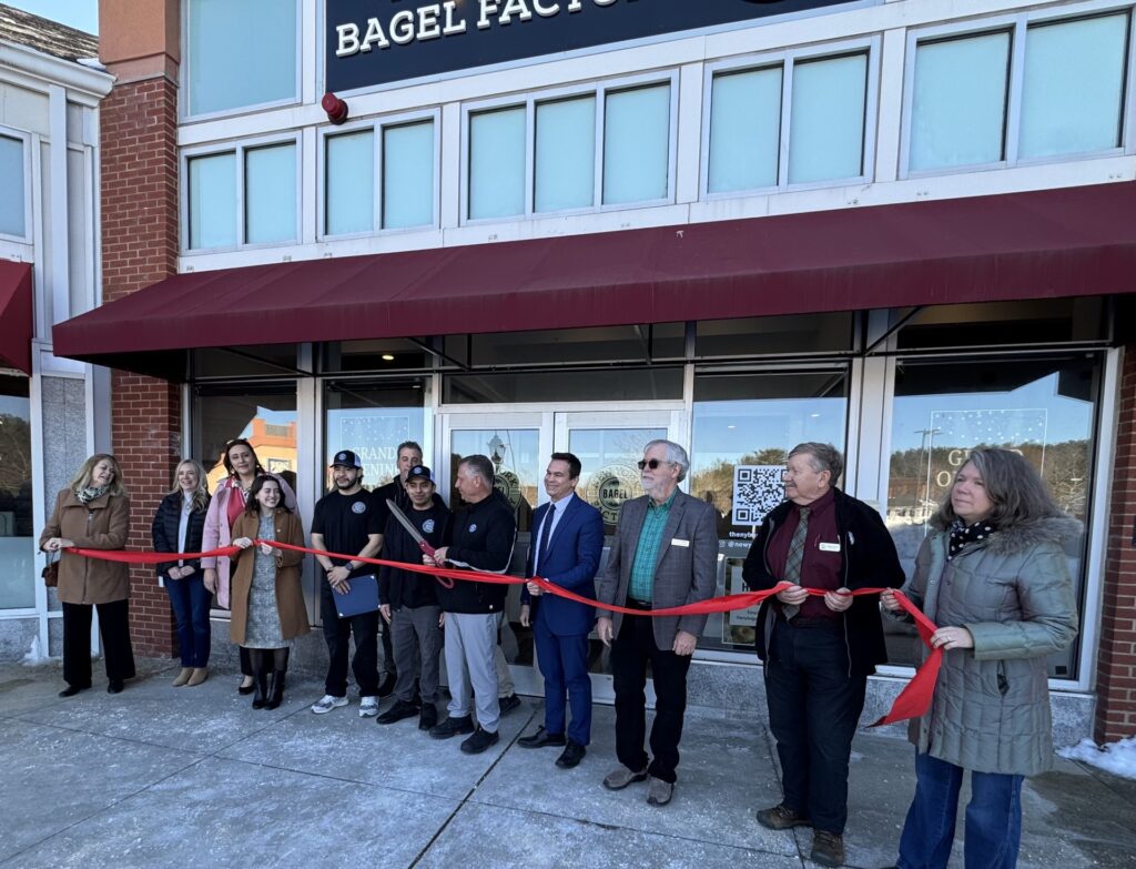 A line of people holding a red ribbon in front of a store. A man wearing black is about to take a pair of oversize scissors to the ribbon.
