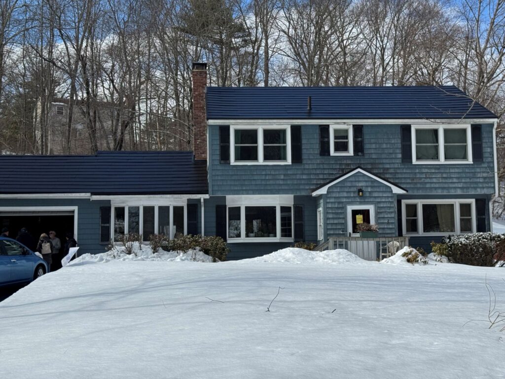 A faded blue garrison colonial house with a slighly shiny black roof. Although there is lots of snow on the ground, the roof is snow-free.