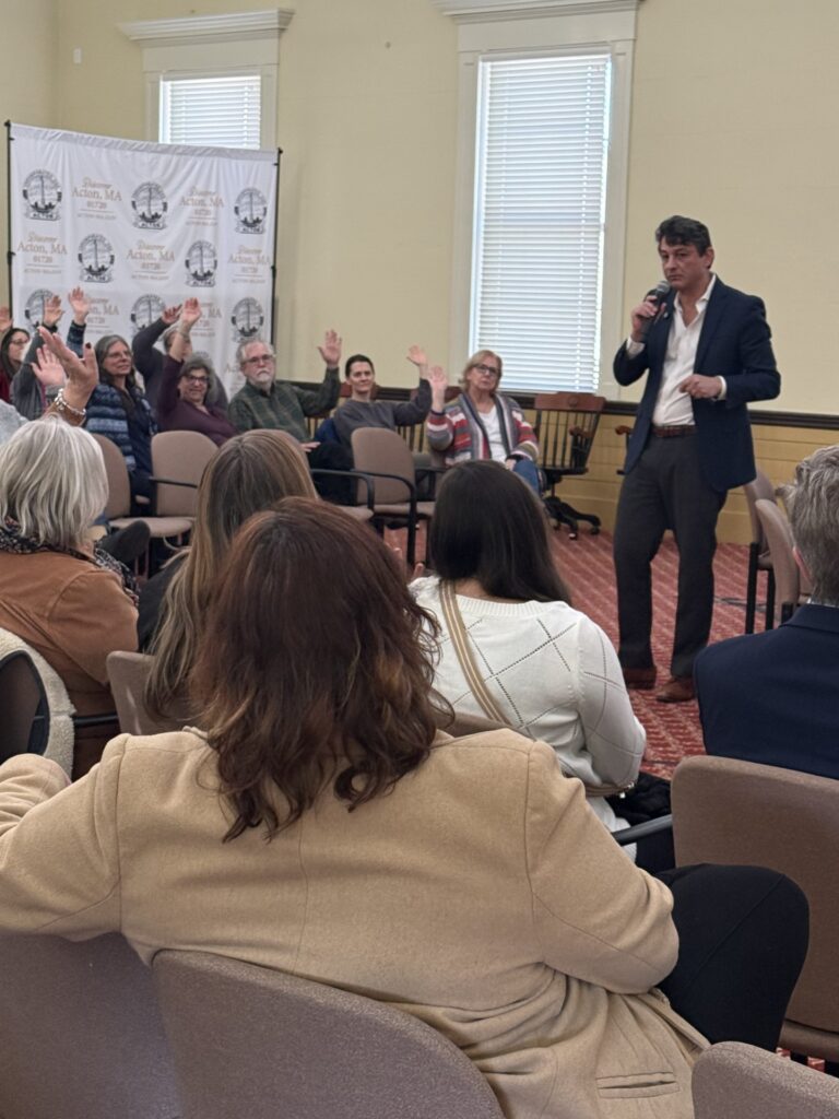 A man in a sport jacket and whit shirt asks the audience a question. In the background, most hands are raised.