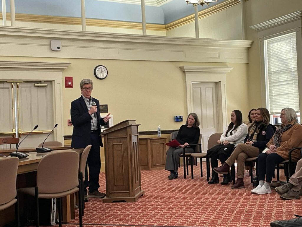 A man in a sport jacket and plaid shirt speaks at a podium.