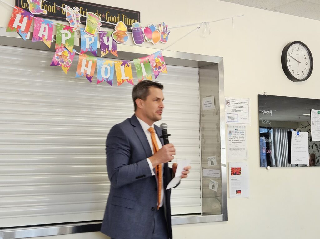 A man wearing a suit, tie, and the beginnings of a beard speaks into a mic while standing under a Happy Holi sign.