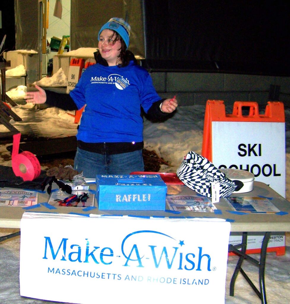 A young woman wearing a Make-A-Wish t-shirt stands at a table with a Make-A-Wish sign, a raffle box, and other fund raising accoutrements.