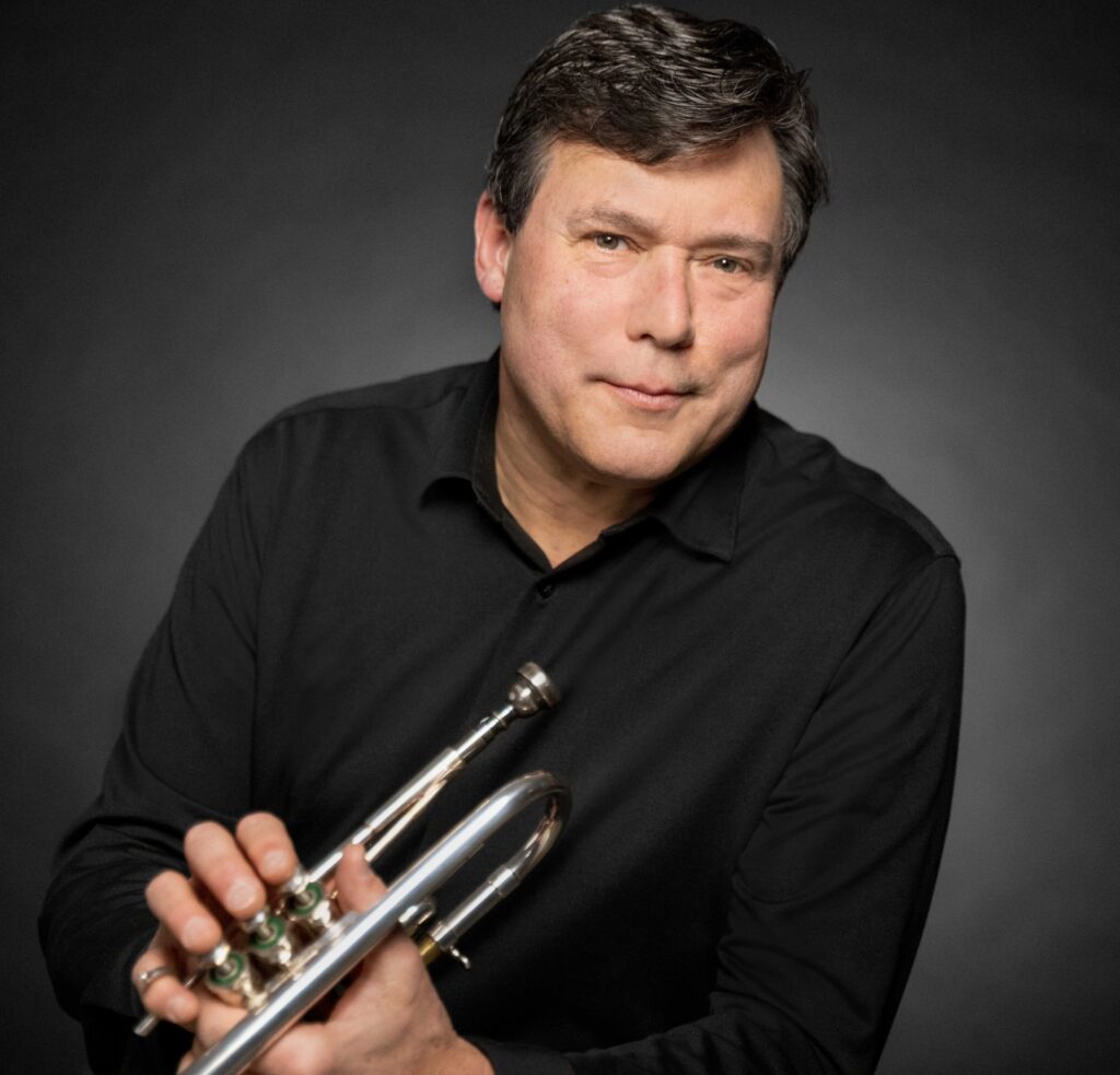 A formal photo of a man wearing a black shirt and holding a trumpet.