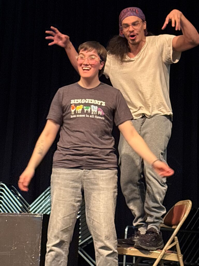 An Afro-Indiginous man with long brown hair and glasses wearing a white tshirt and gray pants stands on a chair with arms raised behind a medium-height, white, transmasculine person with short brown hair, brown eyes and glasses singing.