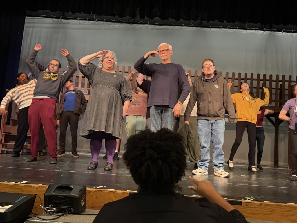 Two people stand at the front of the stage, saluting. Behing them, a number of people are dancing with their hands in the air. In the foreground, a person with fluffy black hair has her hand up facing the stage.