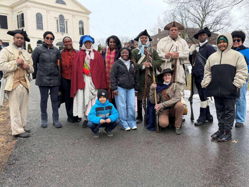 A group of people, most of whom are Black. Many are wearing colonial garb, including a woman wearing a red cape and a man wearing an off-white suit, light brown waistcoat, and a tricorner hat.