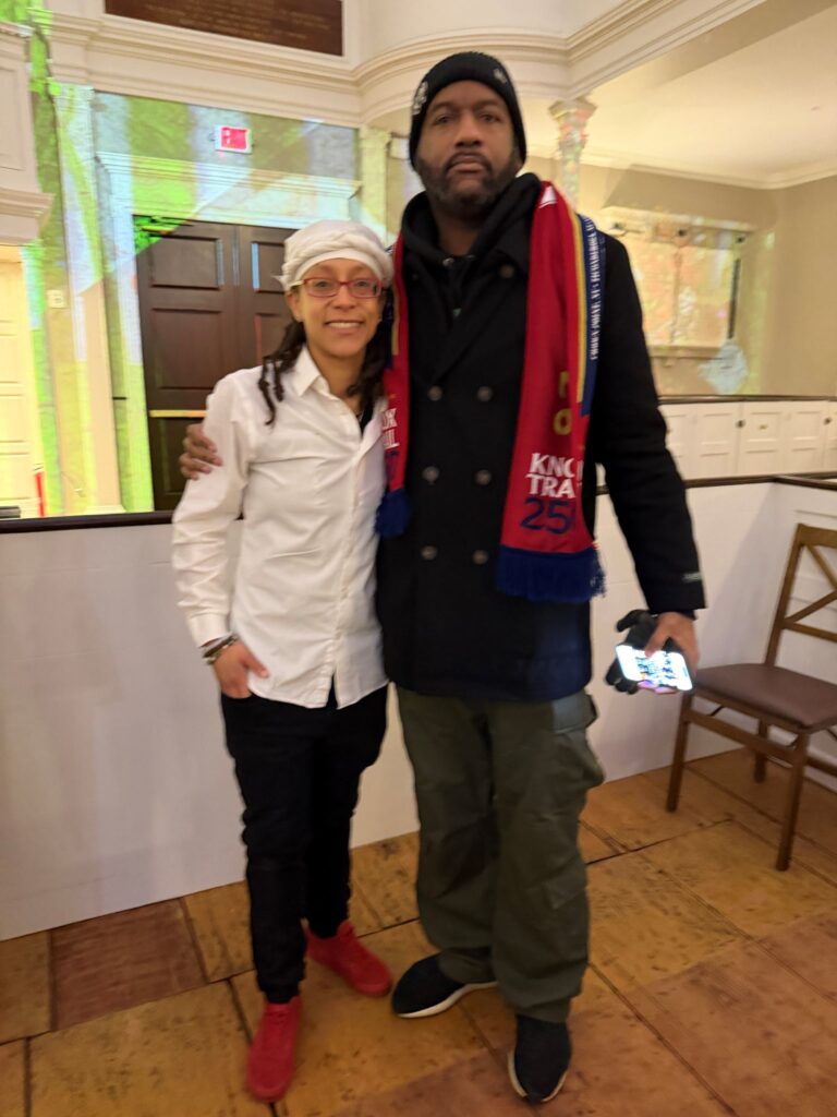 A smiling Black woman wearing a white hat and shirt and red glasses stands next to a much taller Black man who is wearing a black jacket and red Knox Trail 250 scarf.
