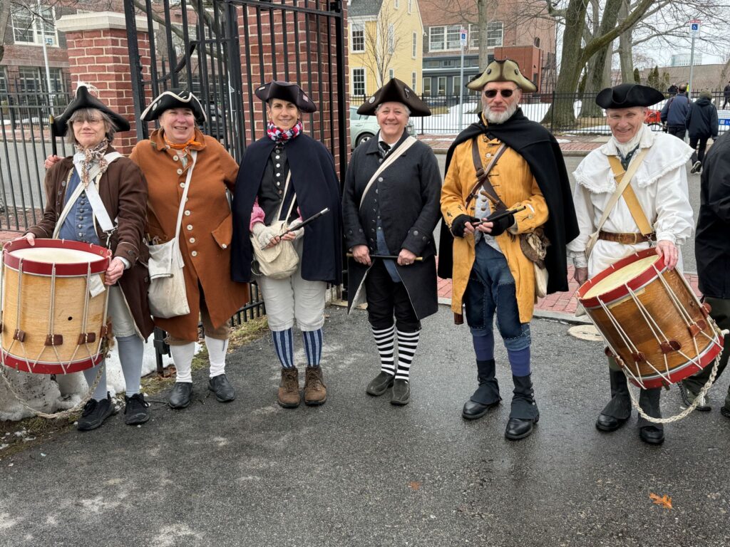 A group of musicians in colnial garb. There are two drummers and four fifers, some of whom are wearing striped socks.