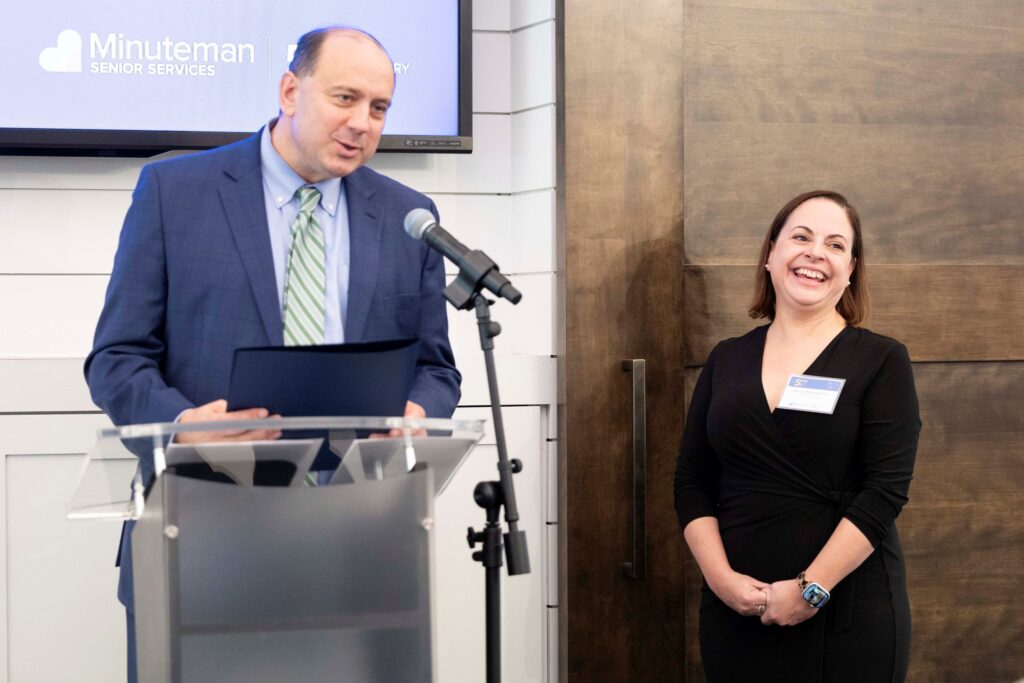 A tall man wearing a suit and tie stands at a podium. A smiling woman in a black dress stands next to him.