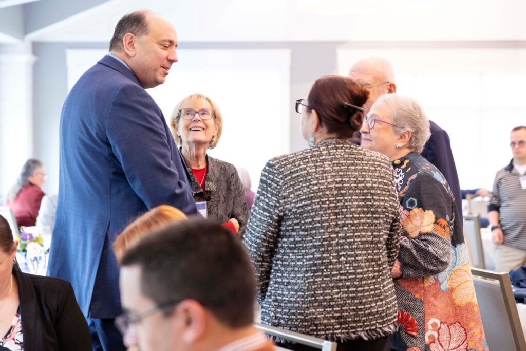 A tall man speaks to a standing group of people, most of whom have gray hair (or not much hair at all).