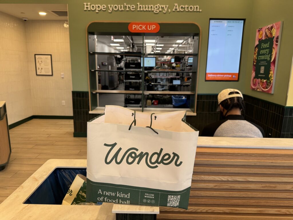 A green and white bag with a Wonder logo sits on a counter. Behind the bag, you can see a person waiting on a bench, and then into an industrial-style kitchen.