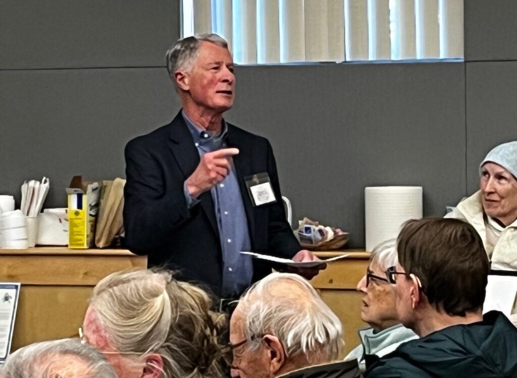 A man in a sportcoat speaks to the crowd. Several heads are in the foreground and a woman wearing a hat is looking at him from the back.