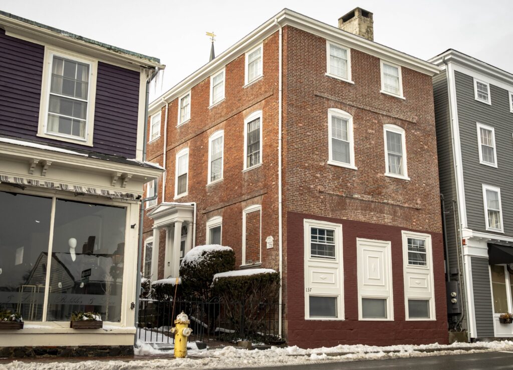 A squarish brick house that is right on the sidewalk. Along the street are several windows; the entrance is on the side.