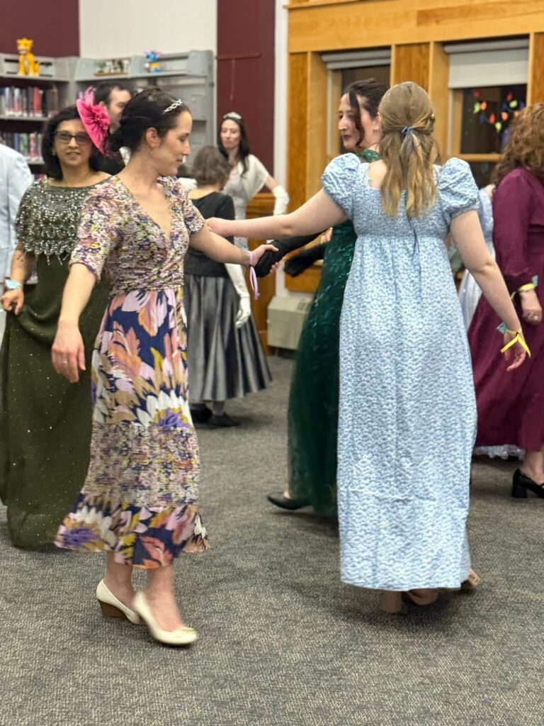 In the foreground, four women are dancing with their left hands in a circle. In the background, you can see several more sets of people dancing.