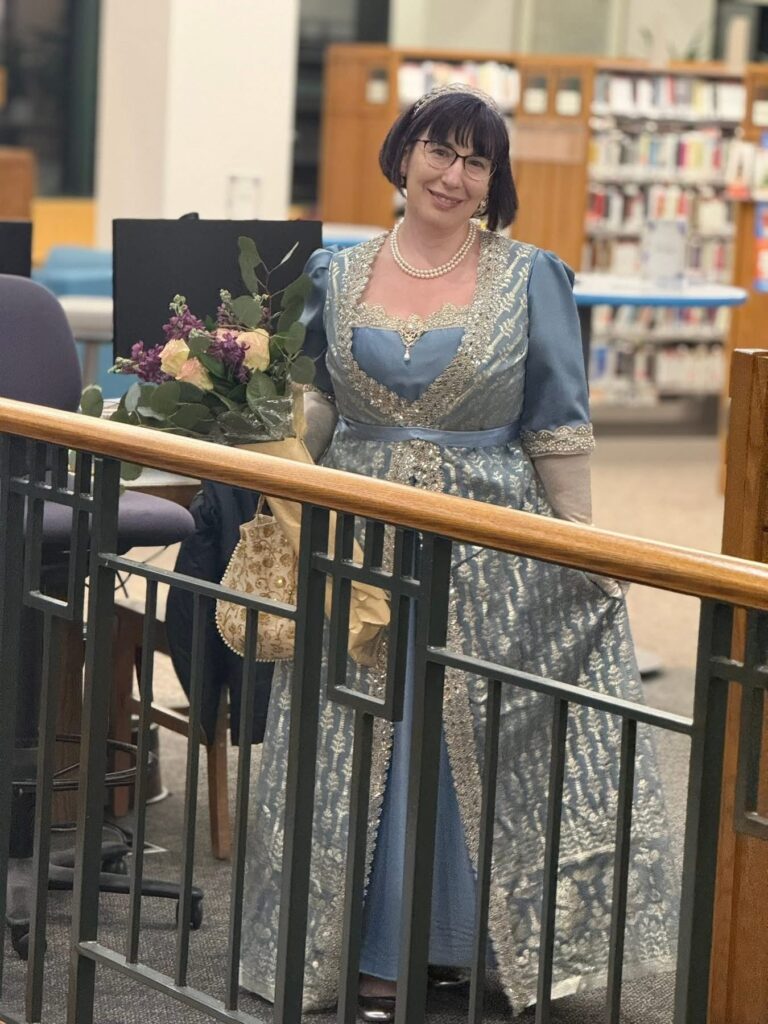 A woman in a blue satin and silver lace gown, wearing full length gloves and a tiara, shows off her lovely handiwork.