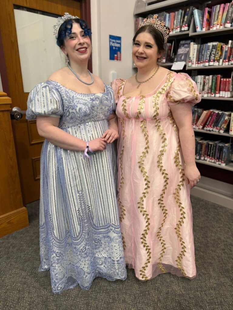 Two woman wearing empire waisted, puff sleeved dresses made with silky fabric. Both women are wearing tiaras to complete their ballroom look.