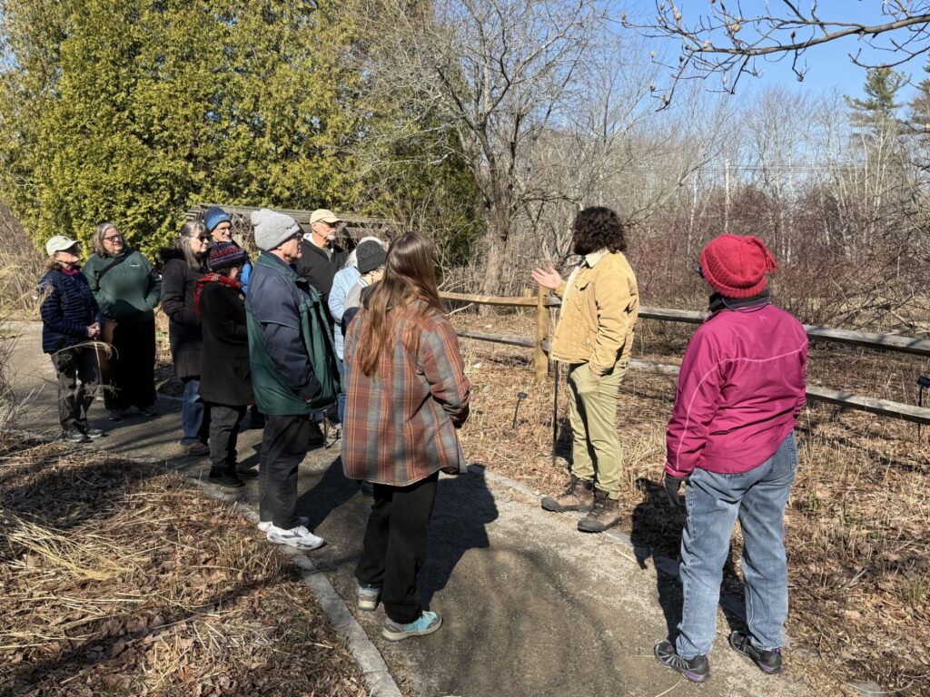 A group of people stand on a walkway next to a wooden fence. A man in a brown jacket is speaking to the group.